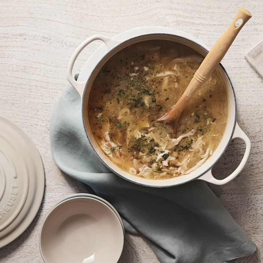 Le Creuset støpejernsgryte med kylling- og urtesuppe, servert med en tre-skje, illustrerer gryten i bruk.