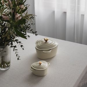 Seasons Love Collection Støpejernsform Hjerte Krem 10cm 0,3L plassert på et bord ved siden av en vase med blomster, illustrerer stilfull bordservering.