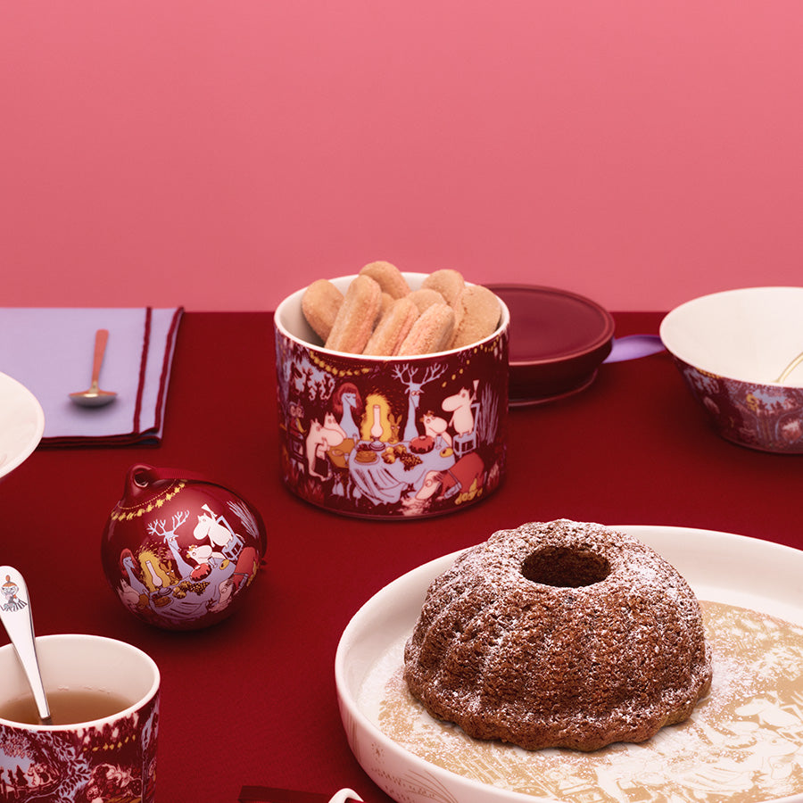 Moomin Arabia krukke på bordet med julekaker og Mummi-figurer, sammen med festlige serveringsretter.