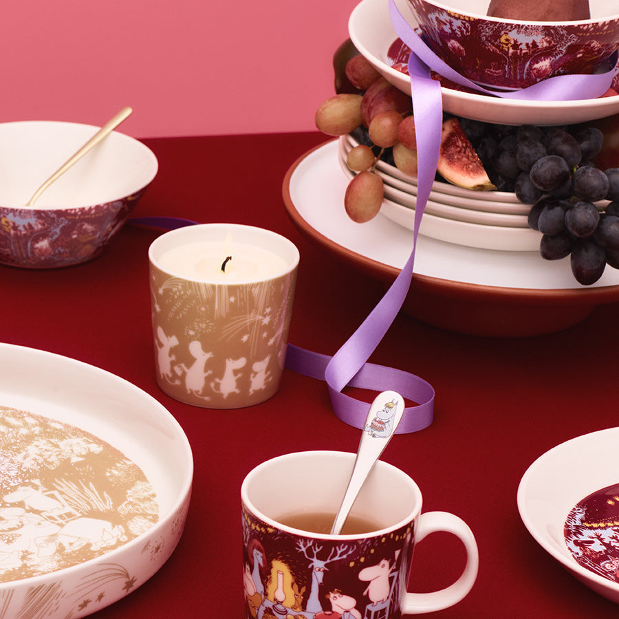 Moomin Arabia Festlige Stunder Mummi stearinlys plassert på et dekkt bord med Mummi-servise og frukt, som skaper en koselig atmosfære for festlige anledninger.