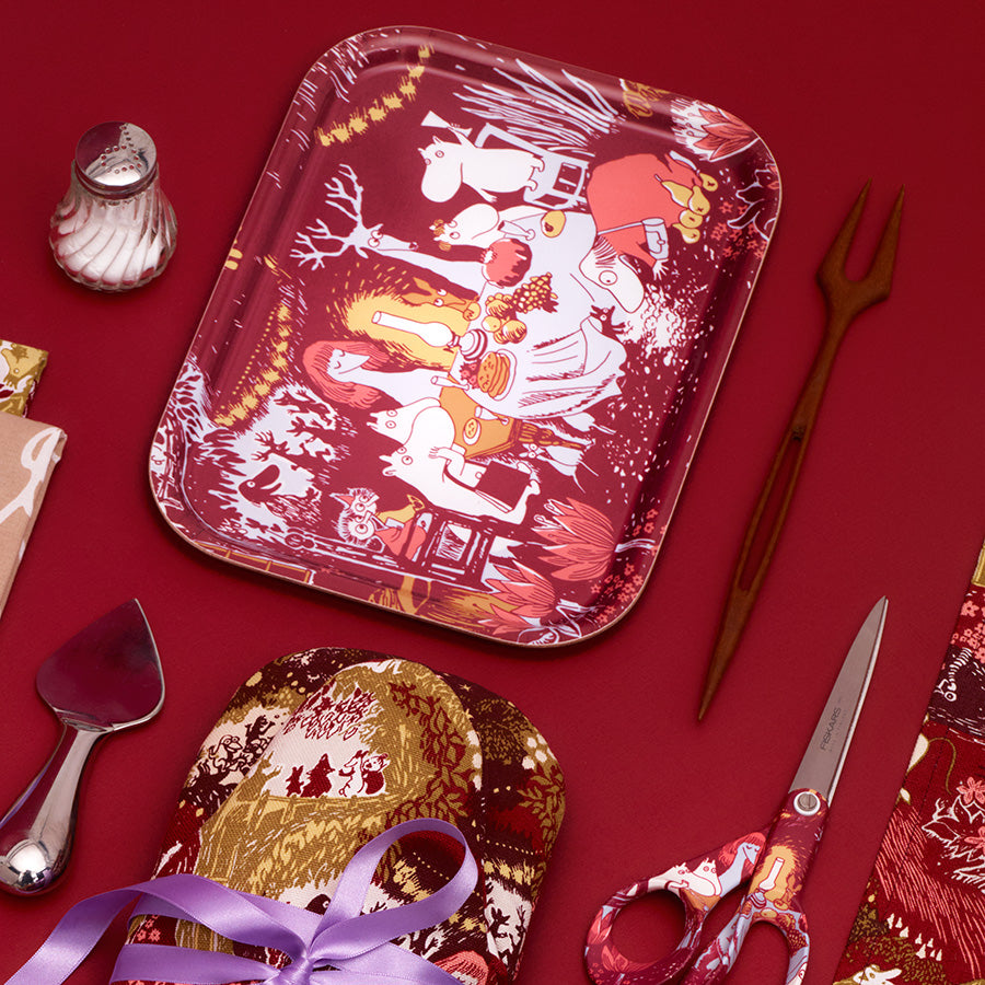 Moomin Arabia serveringsbrett med festlig design, plassert på et rødt underlag med tilbehør som bestikk og servietter. Ideelt for servering av mat og drikke under feiringer.