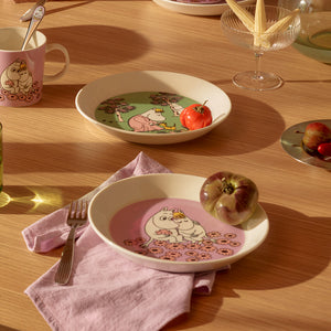 Mummi asjett på et trebord, designet med Mummitrollet og Snorkfrøken, sammen med frukt og servietter, som fremhever et romantisk og nostalgisk måltid.