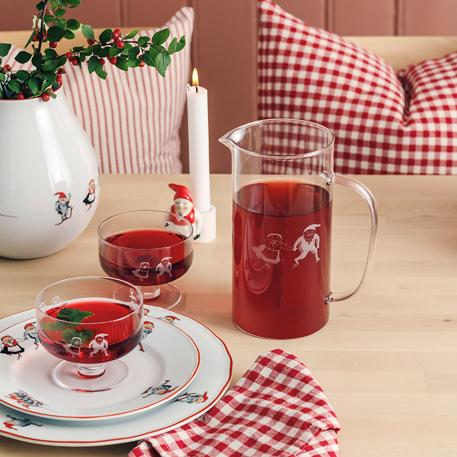 Porsgrund Porselænsfabrik Nisseservise Glassmugge 1,5L på et juledekket bord med røde drikker, omringet av dekorative glass og julepynt.