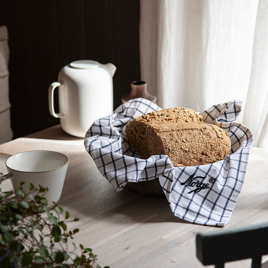 Norgesglasset Kjøkkenhåndkle i blått og hvitt design som dekker brød i en kurv på et trebord, perfekt til kjøkkeninnredning.