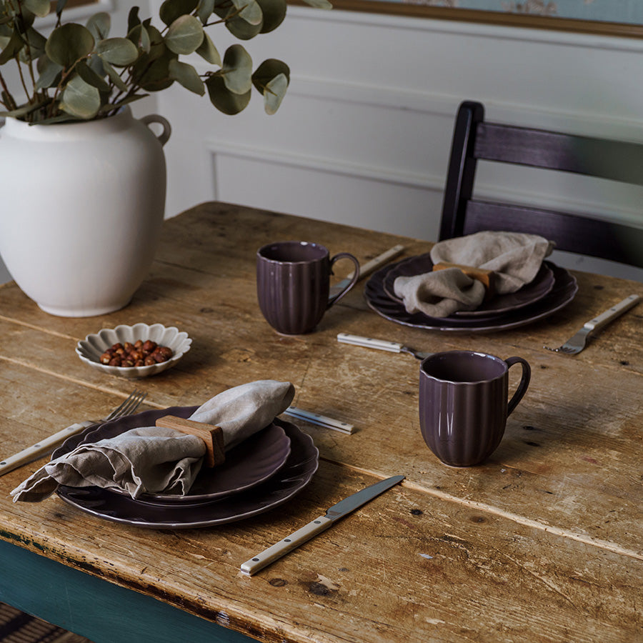 Krus i plommefarge, satt på et rustikt bord med tallerkener, servietter og dekorativ vase, ideell for hverdagsbruk.