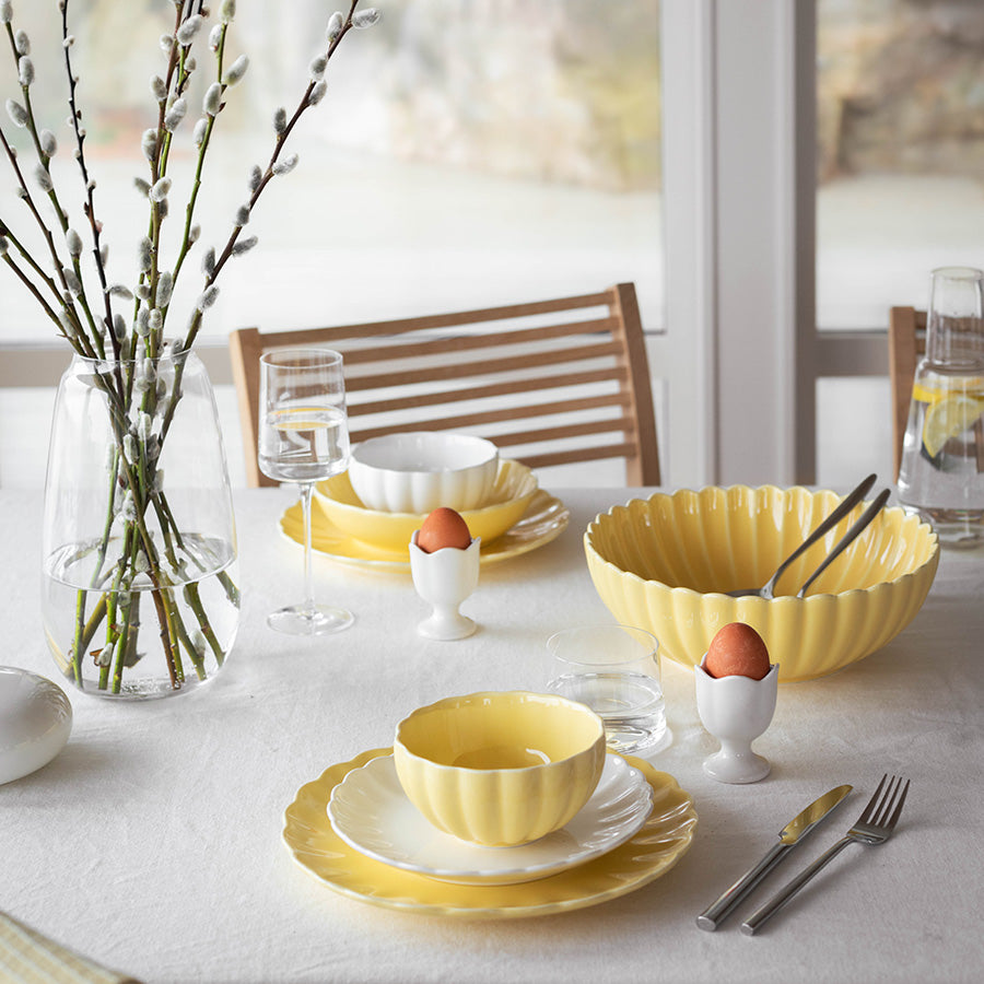 Nova Lilly Frokostservise gul til 2 personer, stilfullt anrettet på bord med gule tallerkener, skåler og glass, skaper en lys og innbydende atmosfære.