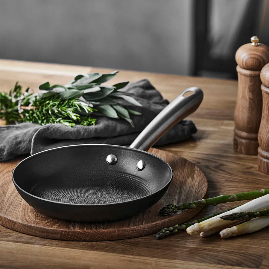 Støpejernspanne fra Pillivuyt Garonne posisjonert på et trebrett med friske urter og annet tilbehør, ideell for matlaging.