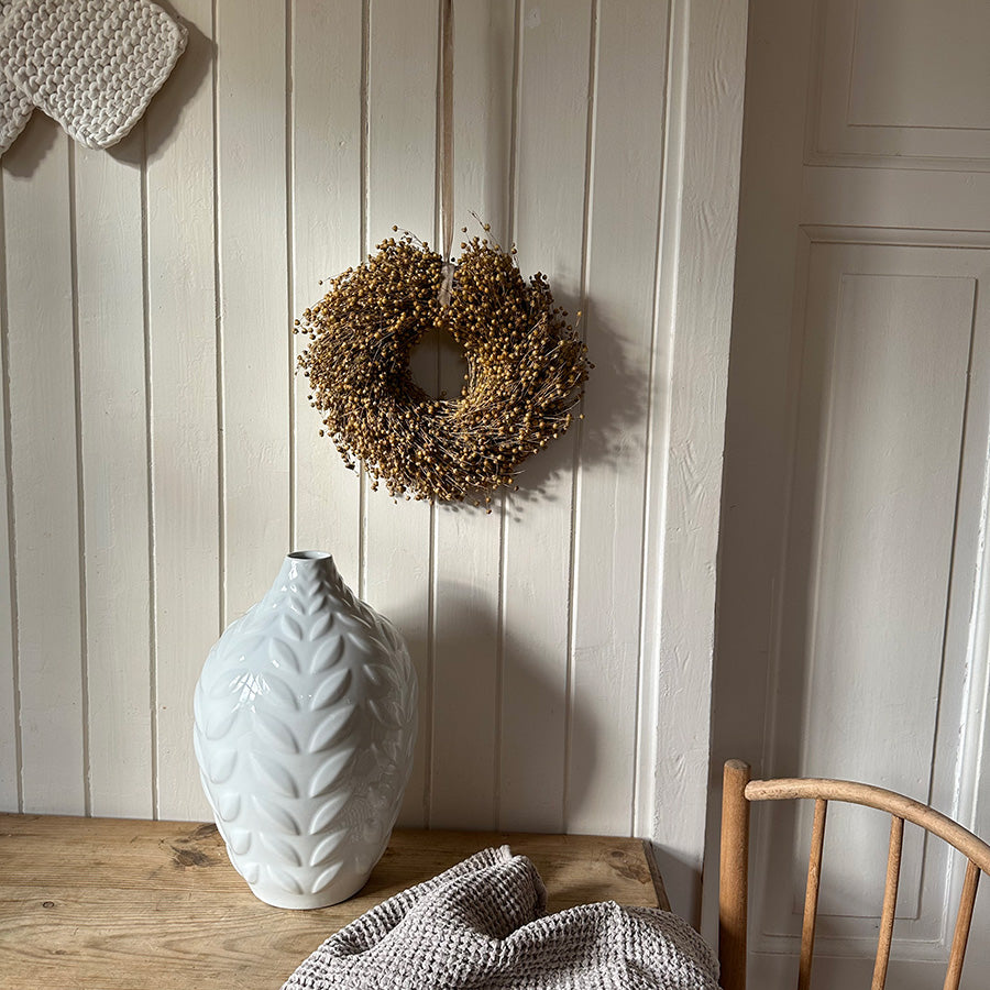 Hvit porselensvase med bladmønster, plassert på trebord, i harmonisk interiør med en krans av tørkede blomster.