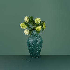 Grønn vase med blomster, designet med bladrelieff, plassert mot en grønn bakgrunn, viser en stilfull kombinasjon av form og farge.