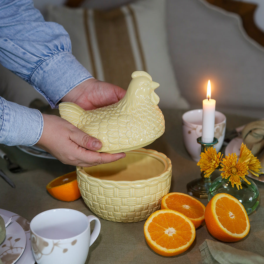 Hånd som holder en gul porselens høne med lokk, plassert over en kurv, i en koselig borddekking med levende lys og appelsiner.
