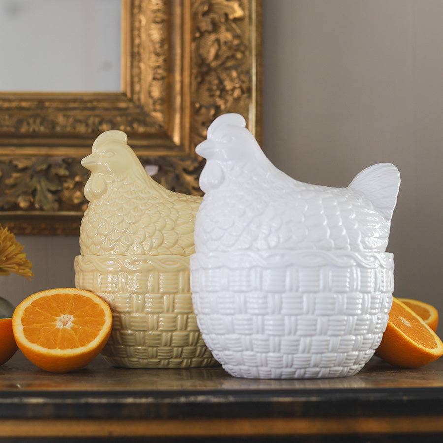 Sammenligning av gul og hvit porselens høne med lokk fra Porsgrunds Porselænsfabrik, begge plassert på et bord med appelsiner og pyntet i nostalgisk stil.