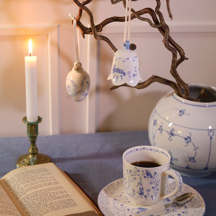 Stilig interiørbilde med blå porselensbjeller hengende på en grein, sammen med en kopp te og en lysestake, som skaper en koselig påskestemning.