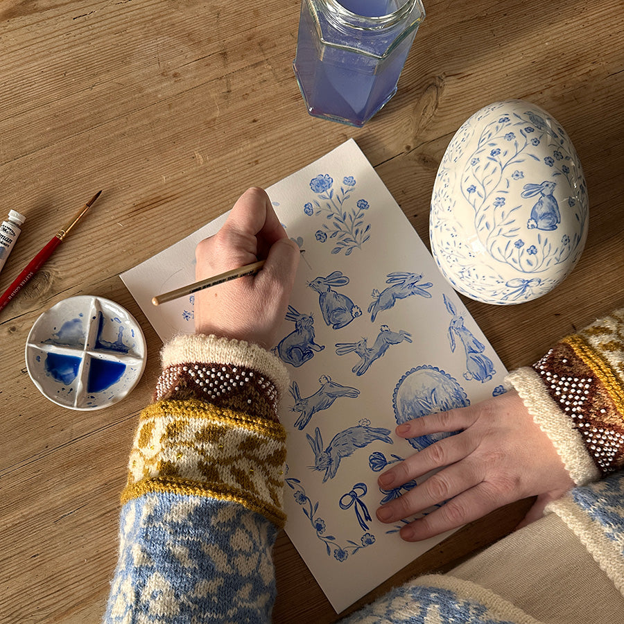 Hånd som maler blå illustrasjoner rundt et porselensegg, viser prosessen av kunstnerisk kreativitet med Vår porselensegg.