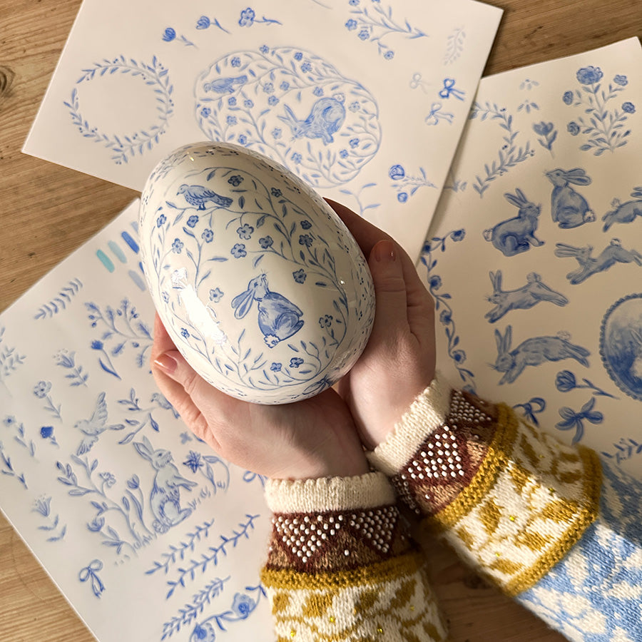 To hender som holder et dekorativt porselensegg med blå blomster og påsketema, omringet av kunstneriske tegninger.