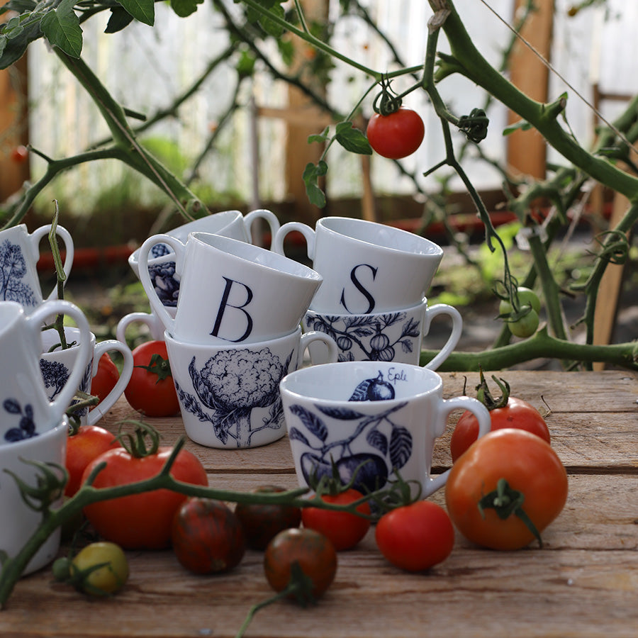 En samling bokstavkrus, inkludert bokstavene B og S, plassert blant tomater i en hage, perfekt for personlig bruk eller som gave.