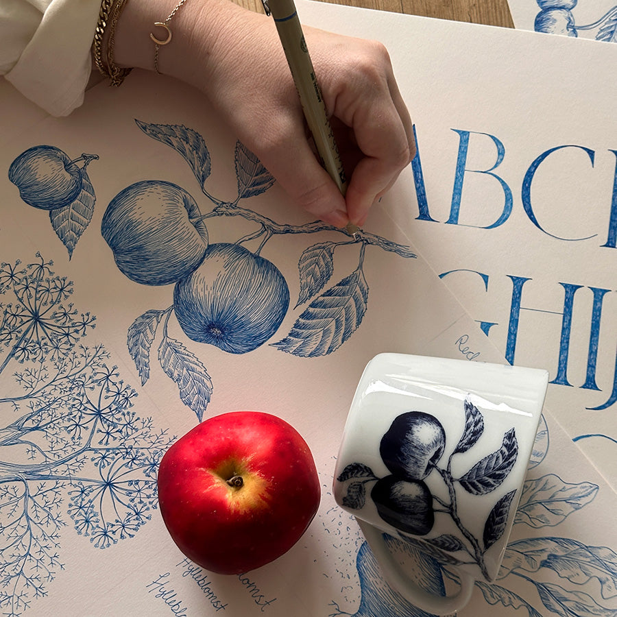 Person som tegner botanisk illustrasjon som inspirerer bokstavkruset, med epler og krus i fokus, kreative prosessen bak bokstavkruset.