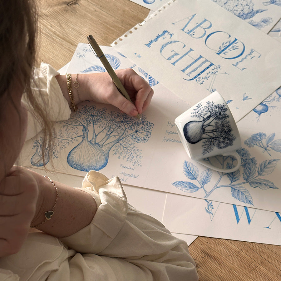 Porsgrunds Porselænsfabrik Bokstavkrus Krus 30cl F, en kunstner i arbeid med kruset i hånden, som illustrerer den botaniske tematikken bak bokstavkrusene.