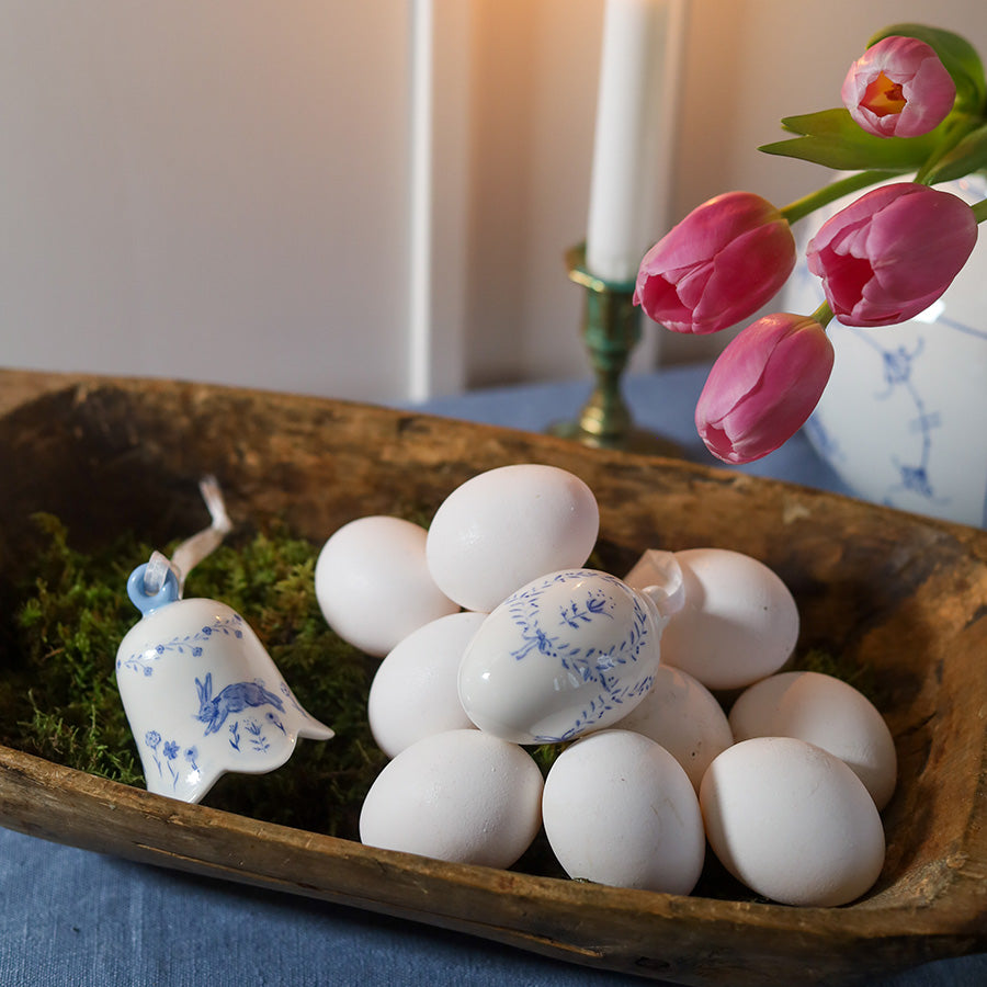 Dekorativ samling med vår porselensbjelle og hvite påskeegg i en naturlig trebolle, omgitt av vårblomster.
