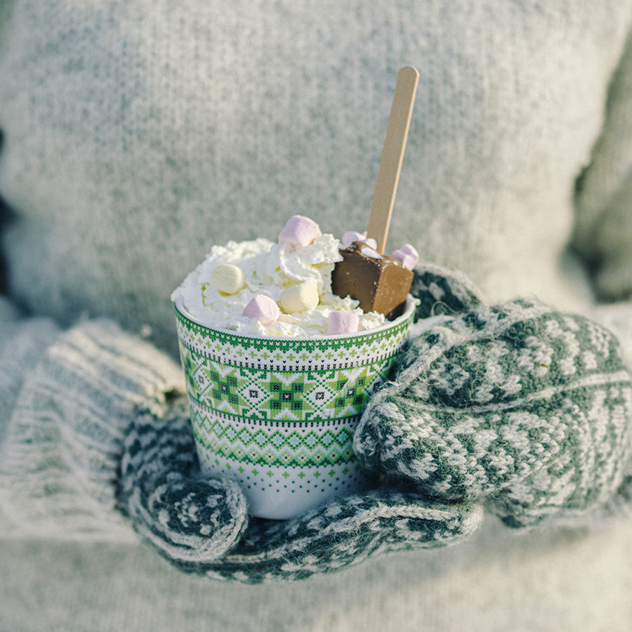 Person iført en grå genser holder et Ingrid krus fylt med kakao og marshmallows, ideelt for varme drikker.