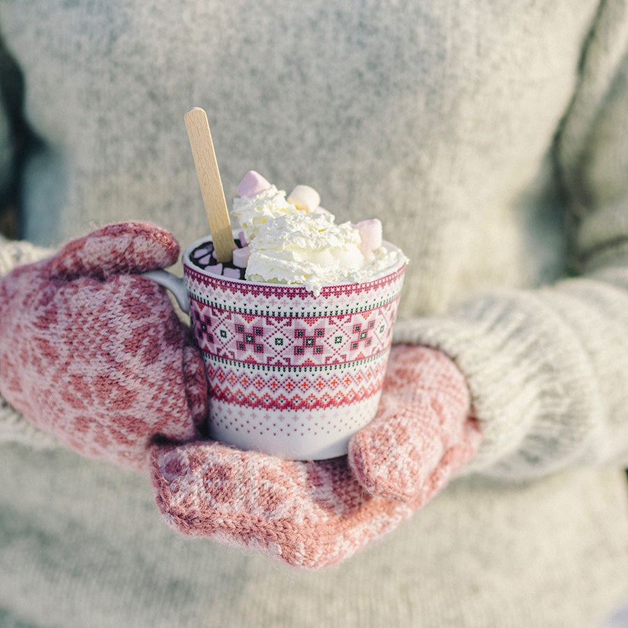 Person med varme og moteriktige votter holder et krus med bunad-mønster, fylt med kakao og pyntet med marshmallows.