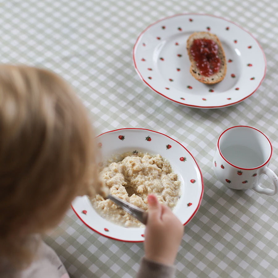 Et barn bruker Porsgrunds Porselænsfabrik barneservise til frokost, med en dyp tallerken med grøt, brød med syltetøy på en flat tallerken, og et krus ved siden av, begge dekorert med nostalgiske markjordbær.