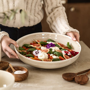 Sjarmerende serveringsbolle i lattefarge, holdes av en person med en frisk salat og blomster, skaper en innbydende atmosfære.