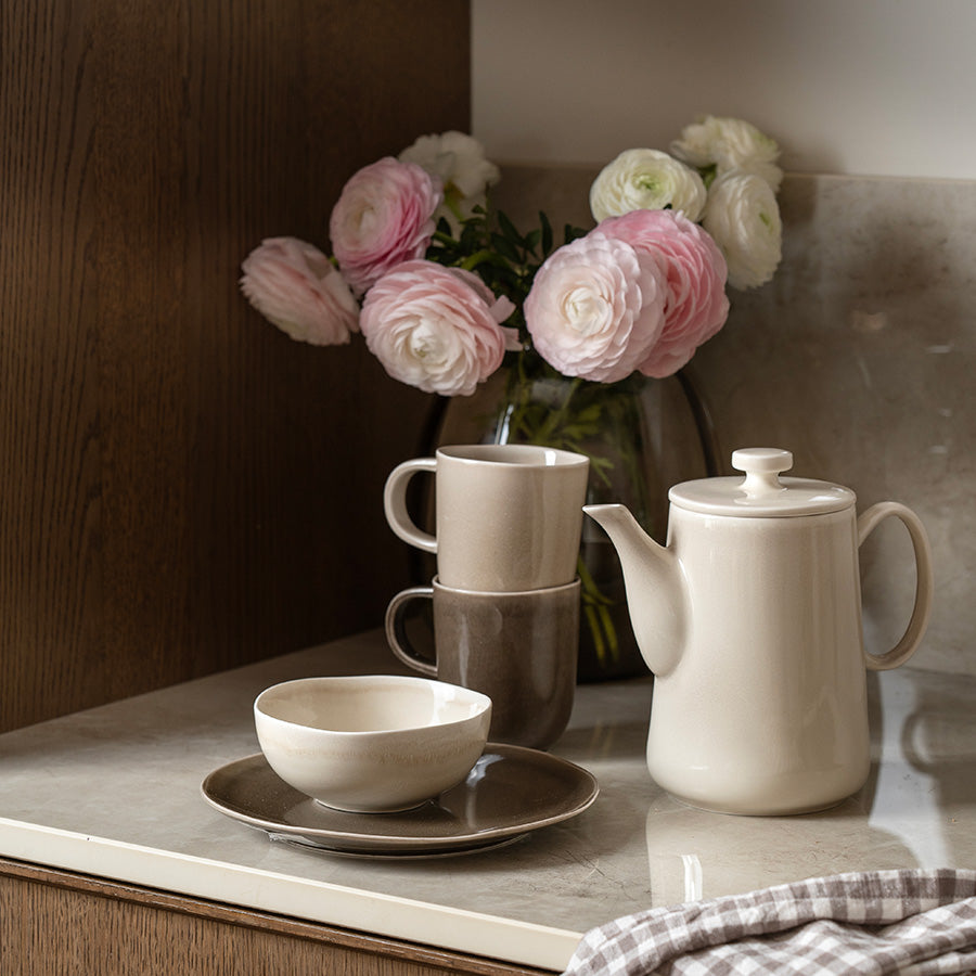 Porsgrunds Porselænsfabrik Ellie tekanne i Latte-farge, plassert på kjøkkenbenk med blomster og kopper, perfekt for en koselig te-stund.