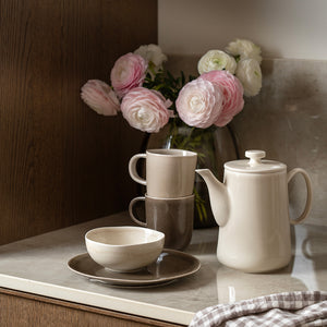Porsgrunds Porselænsfabrik Ellie tekanne i Latte-farge, plassert på kjøkkenbenk med blomster og kopper, perfekt for en koselig te-stund.