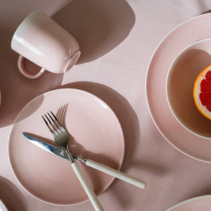 Oversikt over et rosa krus fra Porsgrunds Porselænsfabrik, plassert sammen med en tallerken, bestikk og frukt på en myk rosa bakgrunn.