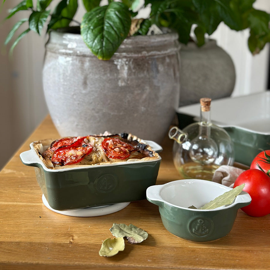 Grønn ildfast form med grateng, porselensskål og kjøkkentilbehør i en lun borddekking