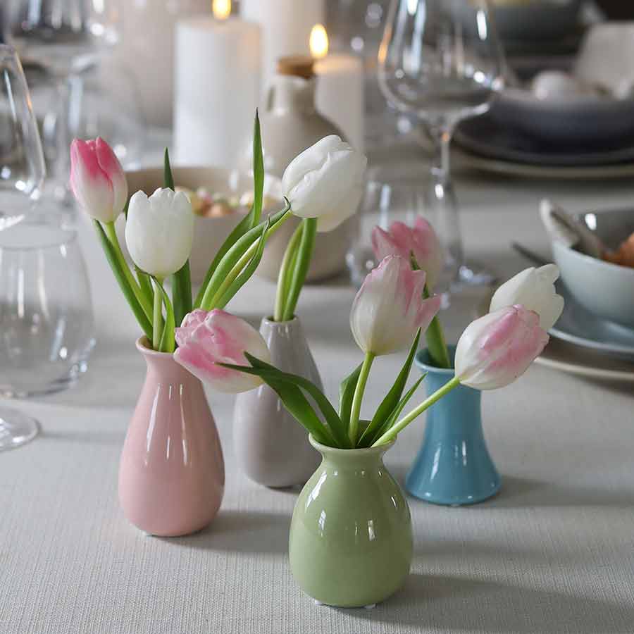 Minivaser med tulipaner på et bord dekket til middag, med stearinlys i bakgrunnen.