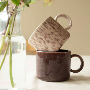 To Porsgrunds Porselænsfabrik Natur Krus, ett i fargen Lyng og ett i en dyp brun nyanse, stablet oppå hverandre på et bord.