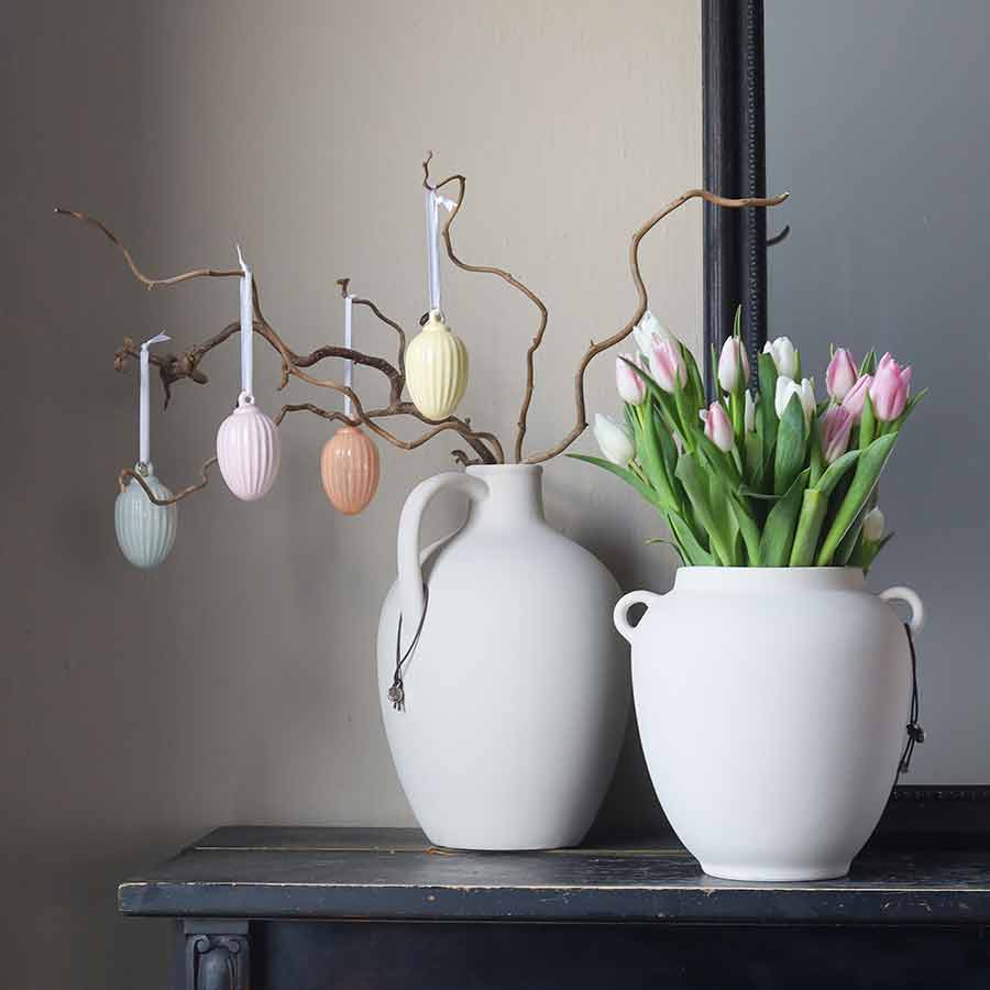 Porselensegg i sart farge, hengt opp på grener og plassert ved siden av en vase med tulipaner, skaper en koselig påskestemning.