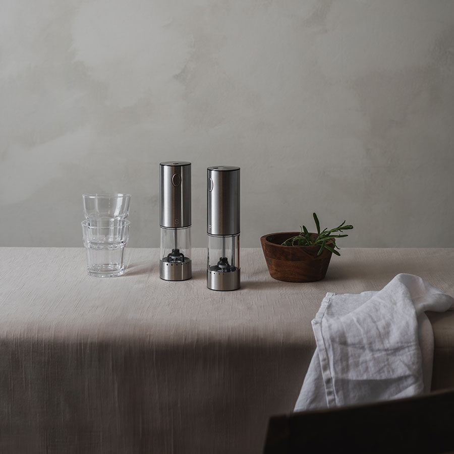 To stilige salt- og pepperkverner i rustfritt stål plassert på et bord med glass og planter, illustrerer praktisk bruk.