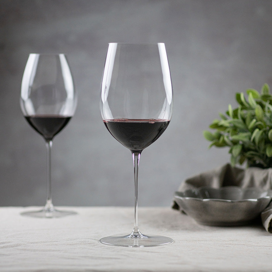 To Riedel Superleggero rødvinsglass med fyldig rødvin, plassert på en bordoverflate, fremhevende elegant design og høykvalitets krystall.