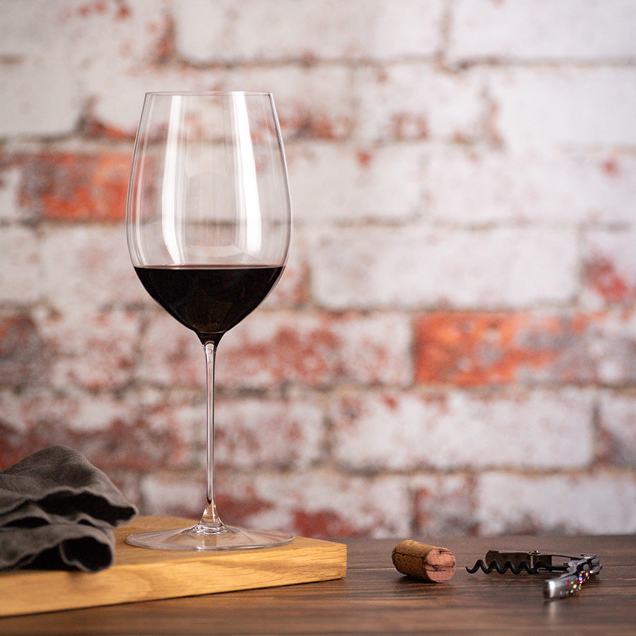 Elegant rødvinsglass fra Riedel, fylt med rødvin, stående på trebrett med kork og vinåpner i bakgrunn.
