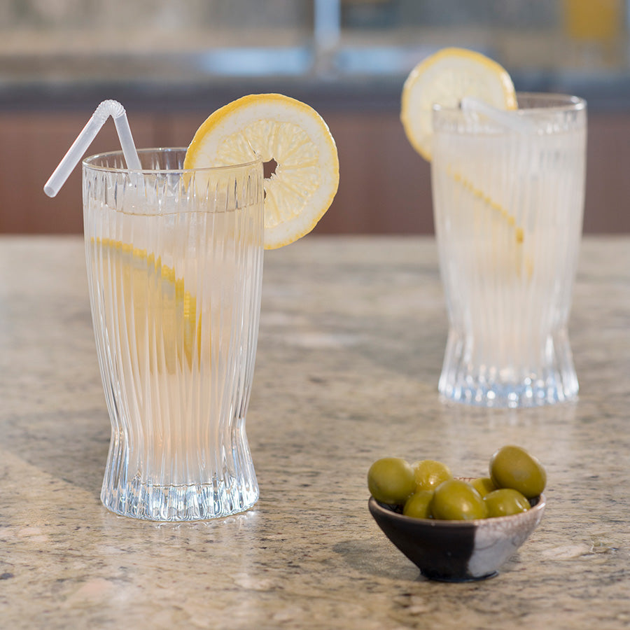 To Riedel longdrinkglass med sitron og sugerør, servert på en lys, steinaktig overflate.