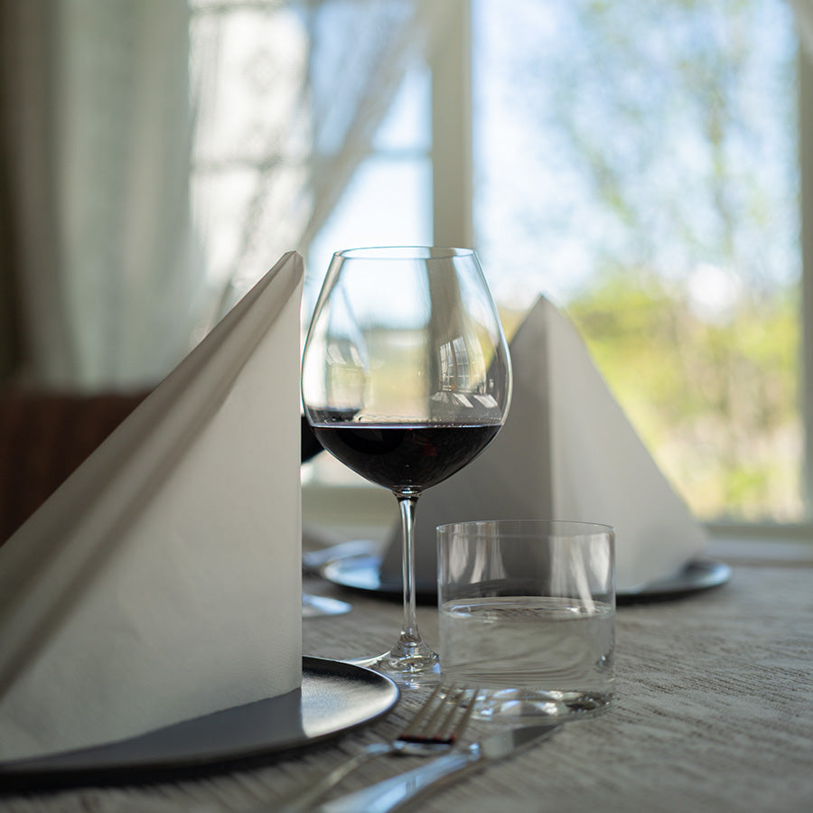 Riedel rødvinsglass med vin i, plassert ved siden av servietter og et glass vann på et elegant dekket bord.