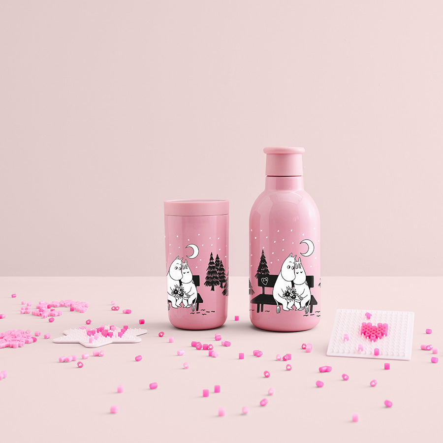 To Stelton Drink-IT Mummi termoflasker i rosa farge, med Mummi-motiver, plassert på et rosa bord med små dekorative elementer.