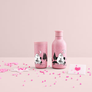 To Stelton Drink-IT Mummi termoflasker i rosa farge, med Mummi-motiver, plassert på et rosa bord med små dekorative elementer.