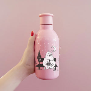 Nærbilde av Stelton Drink-IT Mummi termoflaske 0,5L i rosa, holdt i hånden, med Mummi-figurer og snøfall i bakgrunnen.