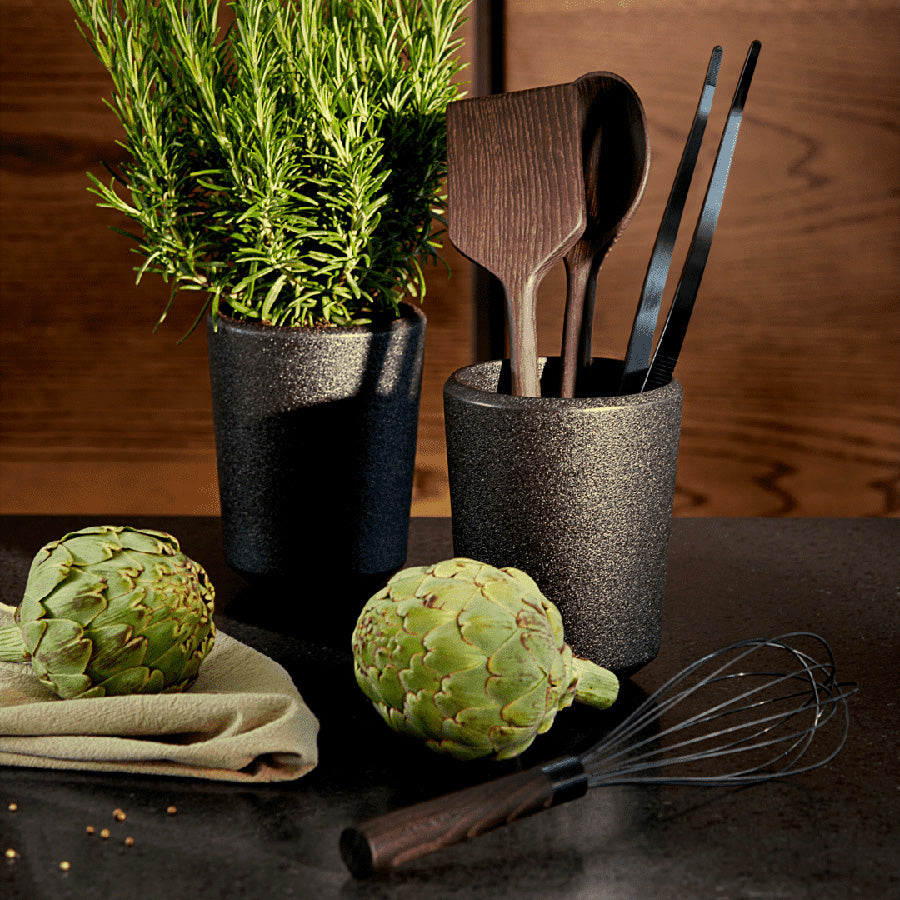 Kolleksjon av kjøkkenredskaper inkludert Rosendahl Rå palettkniv, plassert i en stilren holder med urter og artisjokker.