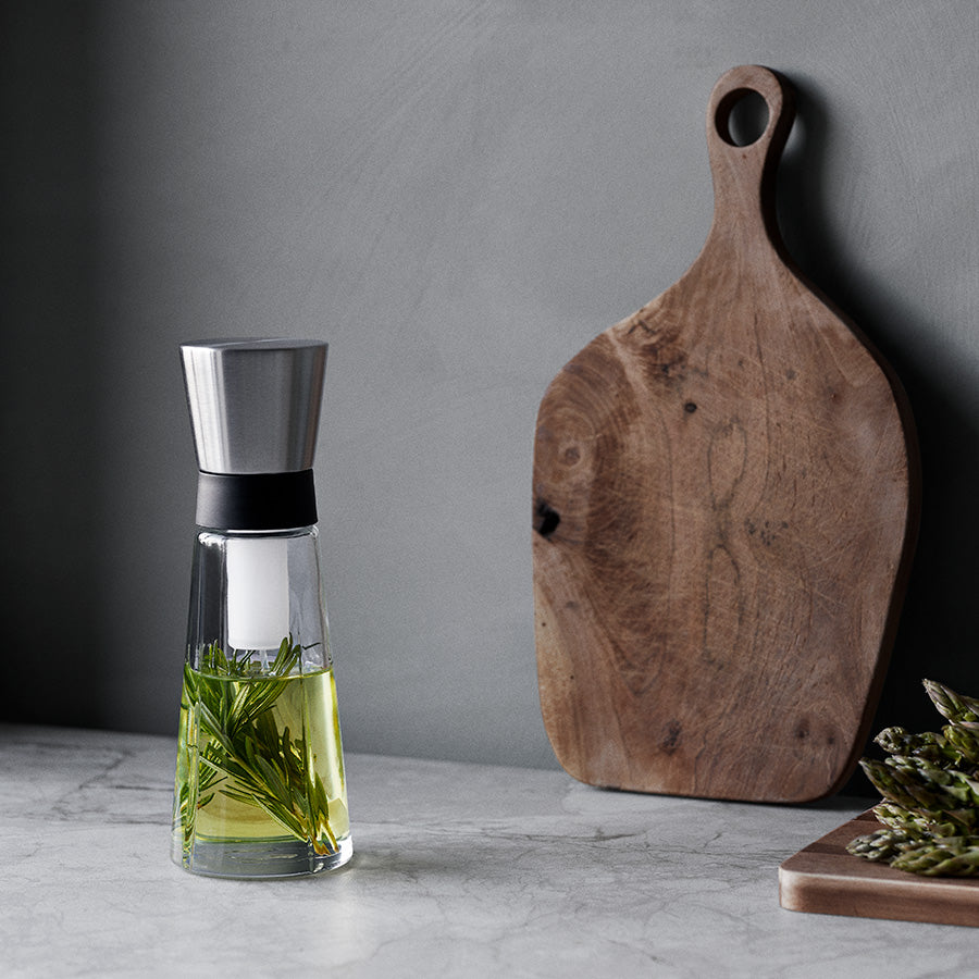 Rosendahl Grand Cru oljeflaske på kjøkkenbenk med urter, sammen med en tre skjærefjøl. Elegant design for optimal bruk.