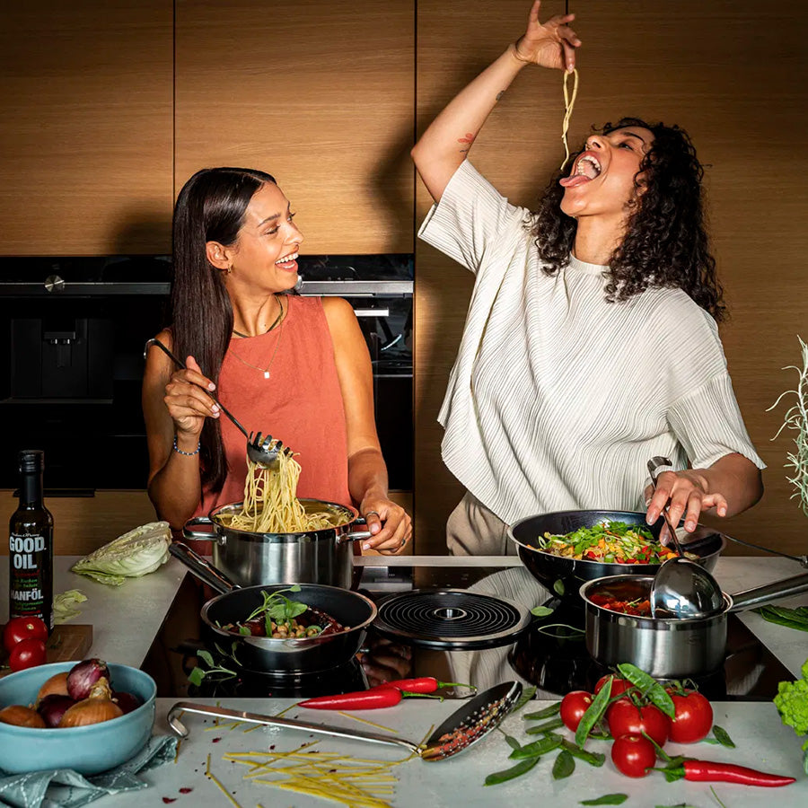 To kvinner lager mat med et Røsle stekepannesett, nyter pasta og grønnsaker i et moderne kjøkken, illustrerer funksjonaliteten til stekepannene.