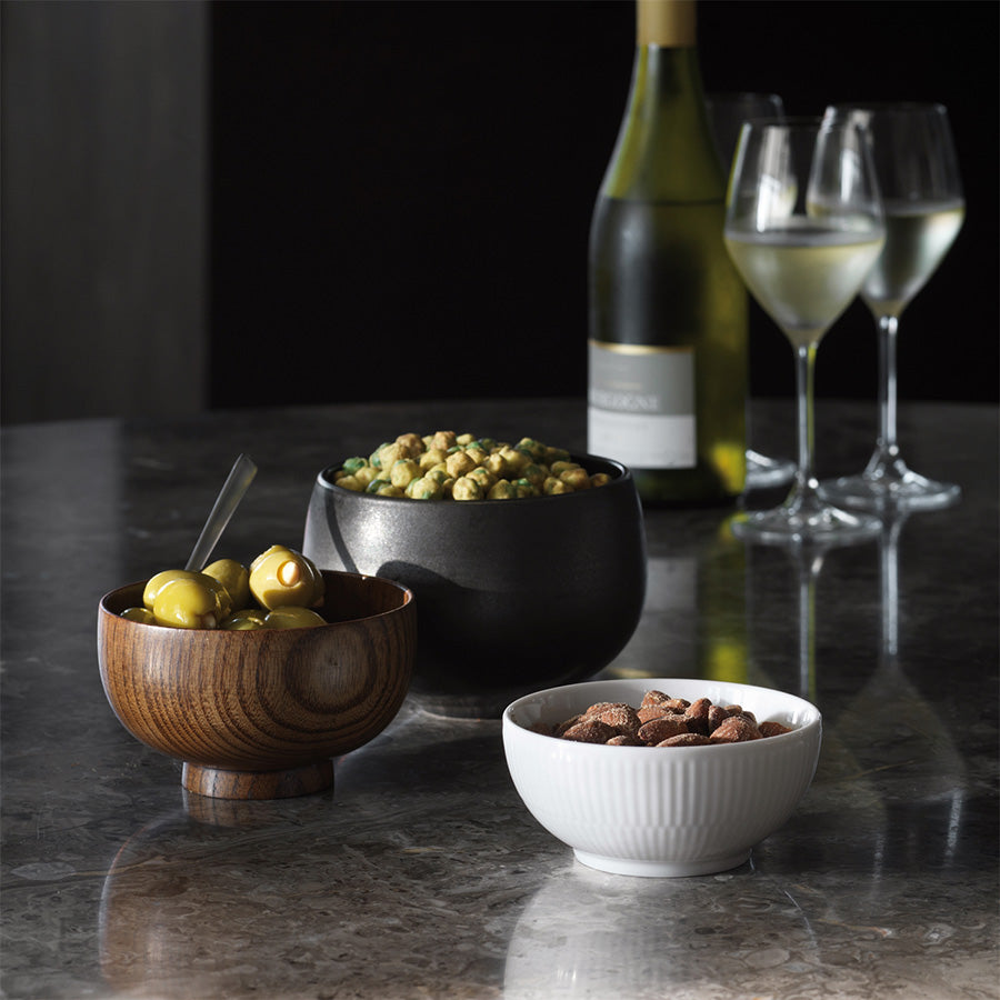 Elegante skåler i ulike materialer på et bord med vin og glass, inkludert en hvit riflet skål fra Royal Copenhagen. Perfekt for snacks som oliven, popcorn og nøtter, og et stilig tillegg til ethvert bord.