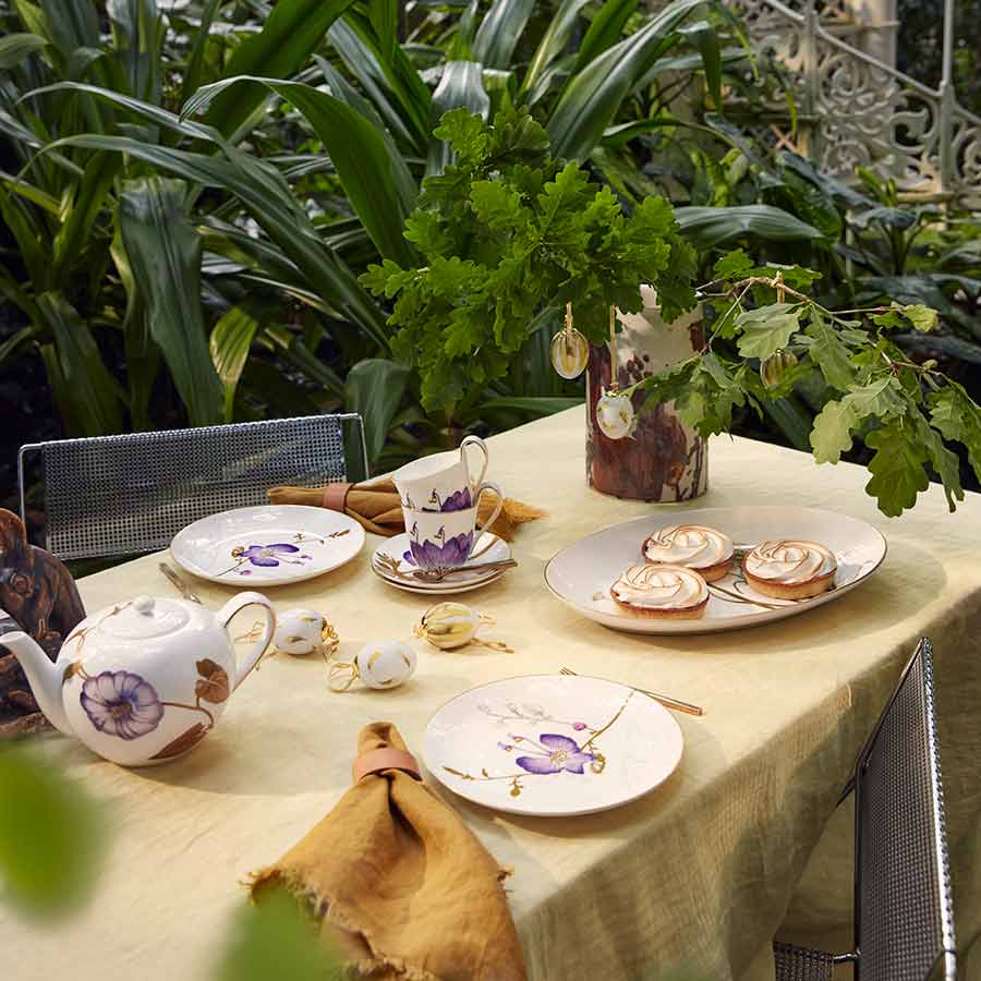 Dekorativt bordoppsett med Royal Copenhagen servise, inkludert tepotte, kopper, og tallerkener, prydet med blomsterdesign. To egg henger på grener i en vase, som tilfører vårstemning til borddekorasjonen.