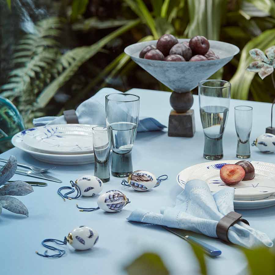 Royal Copenhagen vårkolleksjon 2025 med et stilfullt påskebord. Porselensegg prydet med nøtteskrikefjær sammen med glass og tallerkener på et blått bord.