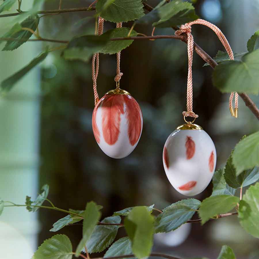 To dekorative egg fra Royal Copenhagen hengende i en grein, prydet med elegante fjærmotiver, ideelle til påskedekor.