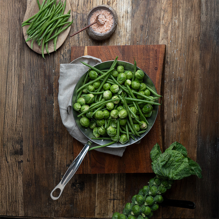 Stekepanne fylt med ferske grønne bønner og rosenkål, plassert på en trebehandling, perfekt for sunn matlaging.