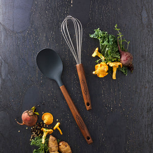 Scanpan Serveringsskje 31cm Karbonisert Ask sammen med en visp og grønnsaker, illustrerer det elegante og funksjonelle kjøkkenutstyret.